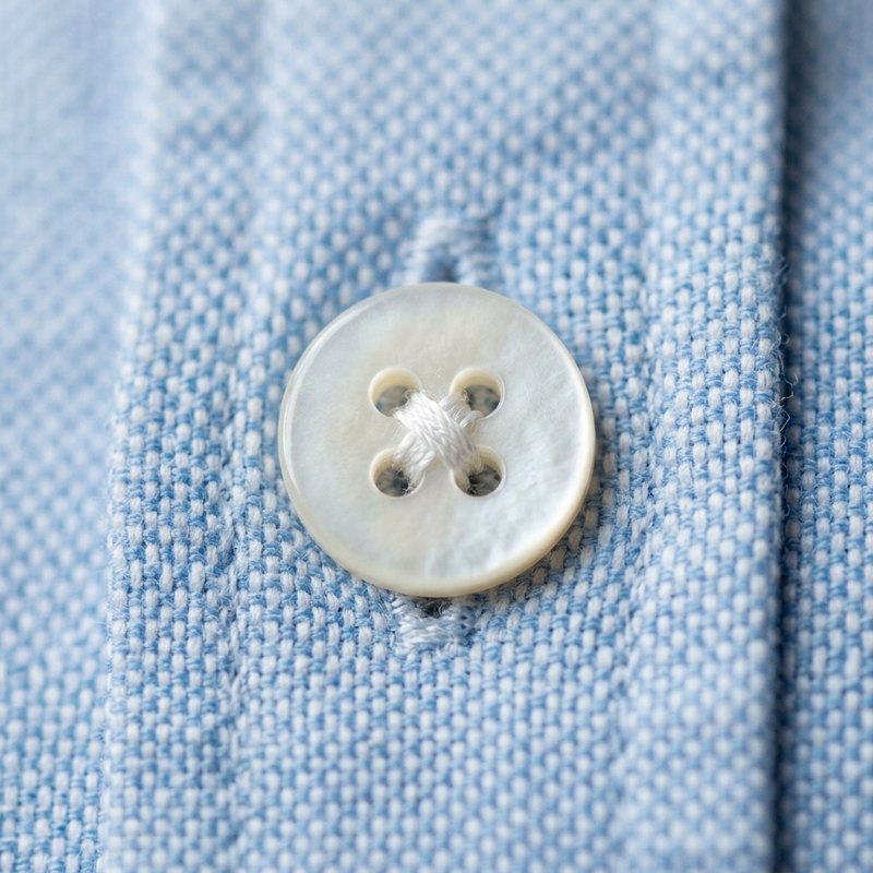 Macro close-up of a neat four-hole shirt button securely stitched onto light blue Oxford fabric
